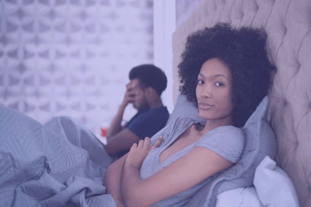 Black couple in bed, main with hand on his face and woman with arms folded looking at the camera