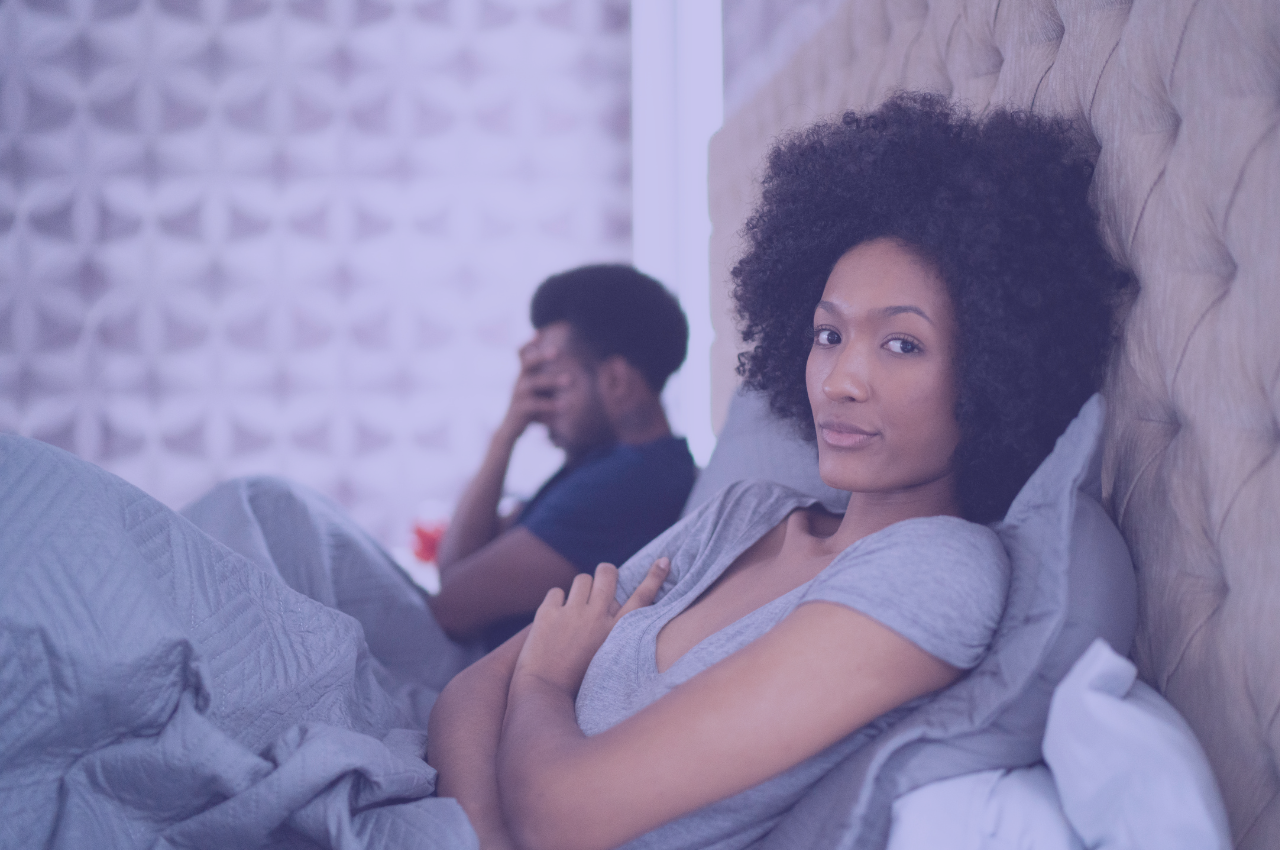 Black couple in bed, main with hand on his face and woman with arms folded looking at the camera